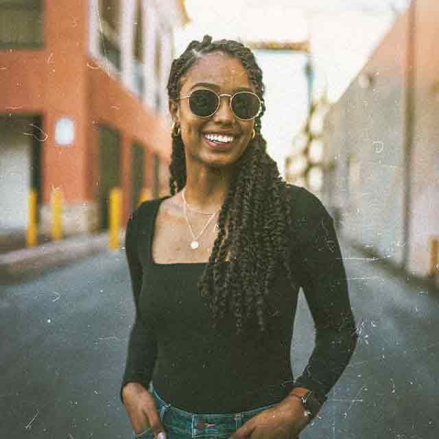 Rochelle standing in the street smiling wearing round sunglasses, black longsleeve tshirt and blue jeans